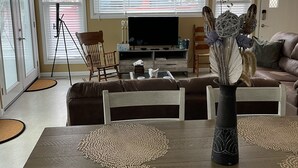 A living room with a dining area, a ceiling fan, a flat-screen TV, and a large window with blinds. - Shore Cliff Cottage  (Ashtabula)