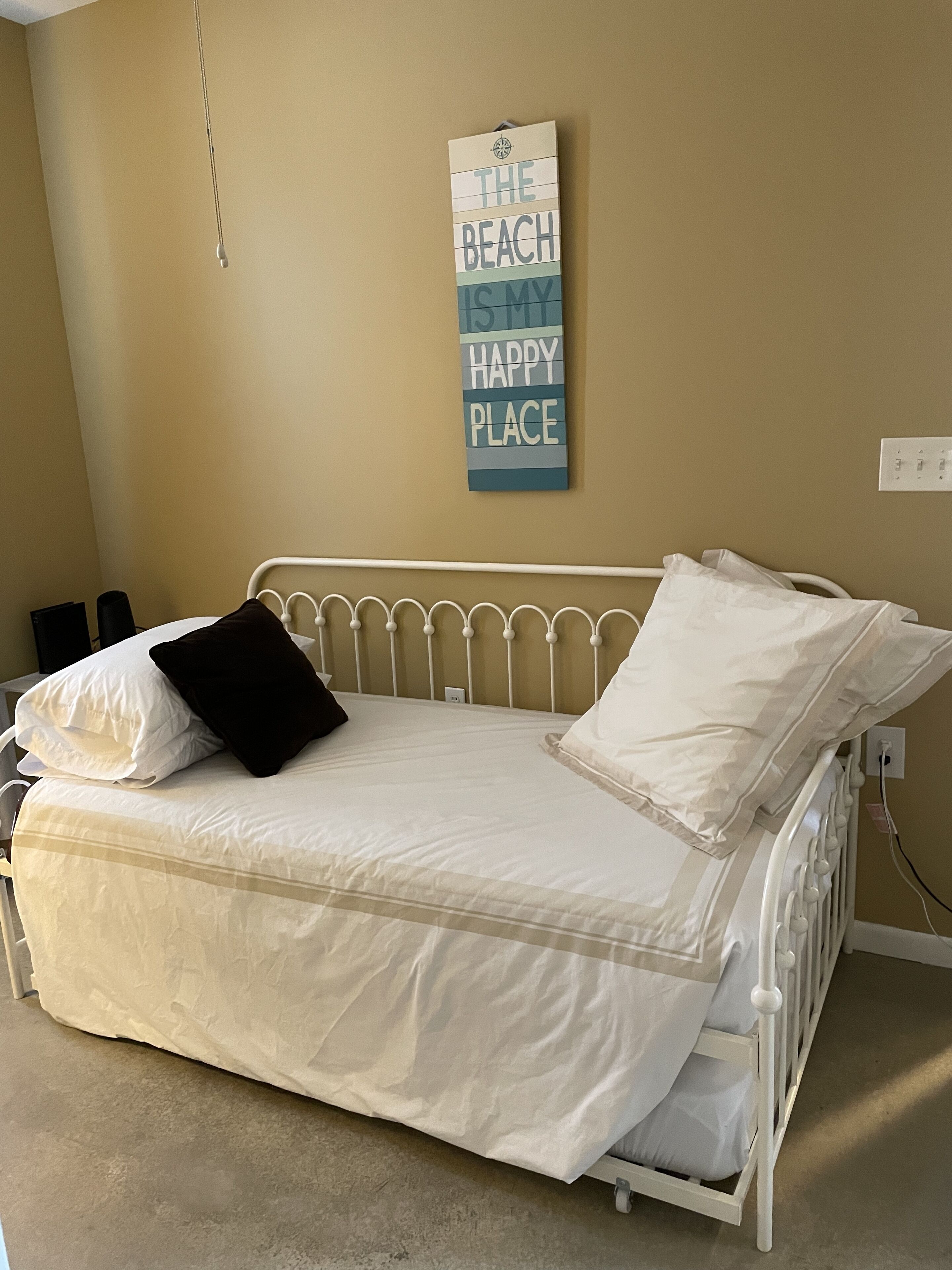 A single-bed room with a white metal frame, beige walls, and a wall-mounted sign that reads 'THE BEACH IS MY HAPPY PLACE.'