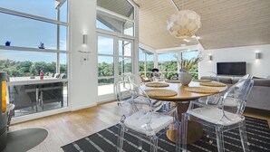A modern living room with large windows, a round dining table with clear chairs, a wooden ceiling, and a TV on the wall. - Stunning home in Hjørring with WiFi (Hjørring)