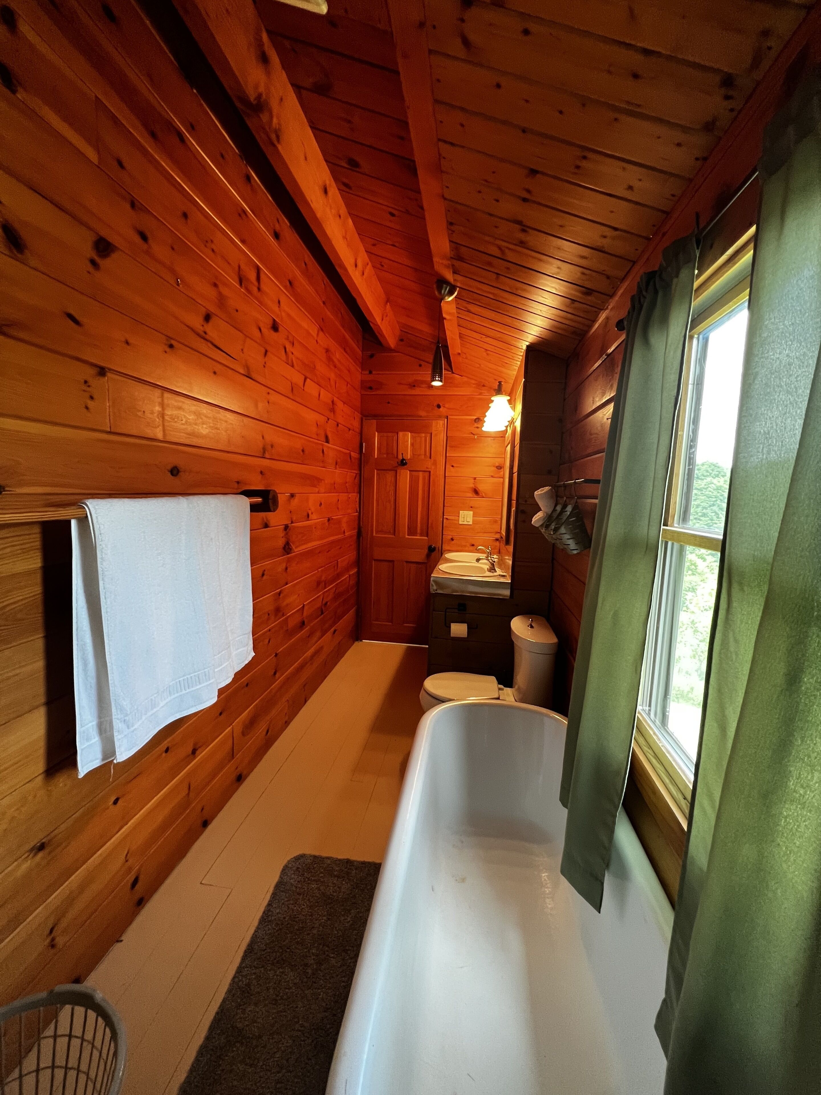 Baño de cabaña de madera con bañera, inodoro, lavamanos y una ventana con cortinas.