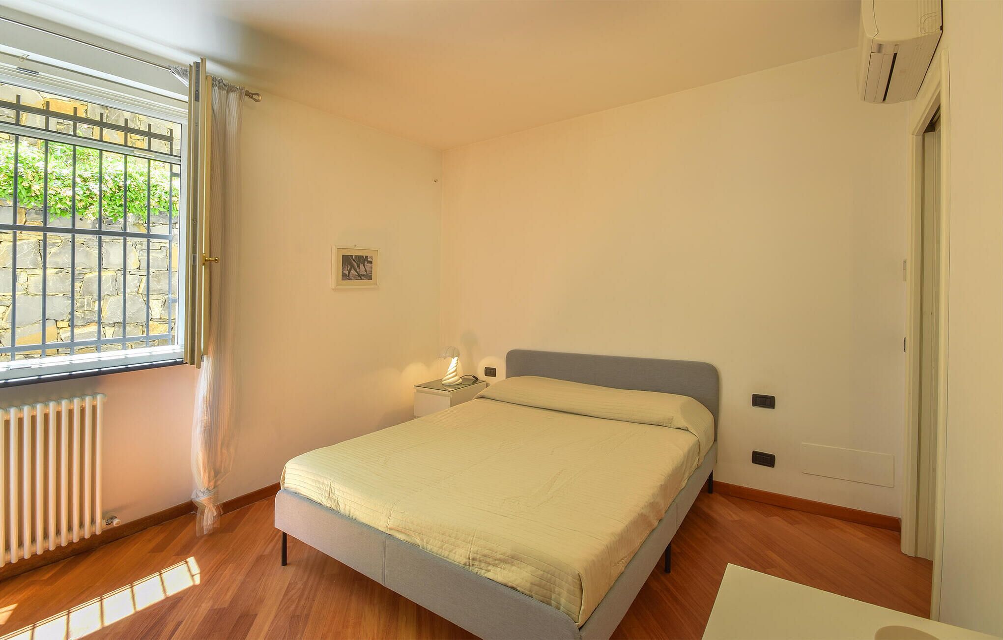 A bedroom with a bed, a nightstand, a window with bars, and a radiator.