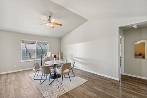 Une pièce spacieuse comprenant une table à manger, des chaises, un ventilateur de plafond et une grande fenêtre avec des rideaux.
