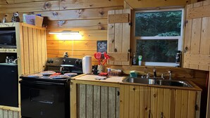 Une cuisine rustique, située dans une cabane en bois, équipée d’un poêle, d’un évier et d’une fenêtre.