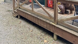Une cabane en bois avec une porte verte, une rampe en bois menant à l’entrée, et deux bannières rouges sur lesquelles on peut lire « BIENVENUE » et « JOYEUX AUTOMNE ».