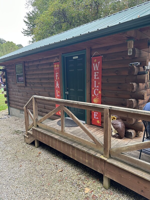 Une cabane en bois avec une porte verte, une rampe en bois menant à l’entrée, et deux bannières rouges sur lesquelles on peut lire « BIENVENUE » et « JOYEUX AUTOMNE ».