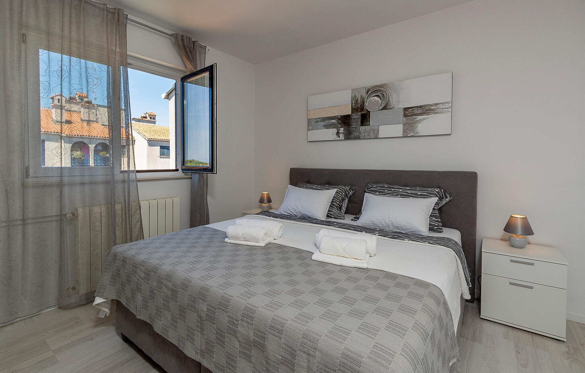A bedroom with a bed, a nightstand, a bedside table, a window, and a painting on the wall.