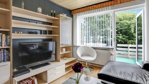 Ein modernes Wohnzimmer mit einem Flachbildfernseher, Bücherregalen, einem weißen Sessel und einem großen Fenster mit Blick auf Bäume.
