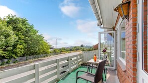 Ein Balkon mit Korbstuhl und Tisch, einer Glastür und Blick auf Grünflächen und Gebäude.