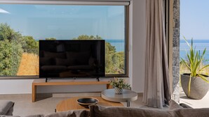 Una sala de estar moderna con un televisor grande sobre un mueble de madera, una mesa redonda y vista al mar a través de un ventanal.