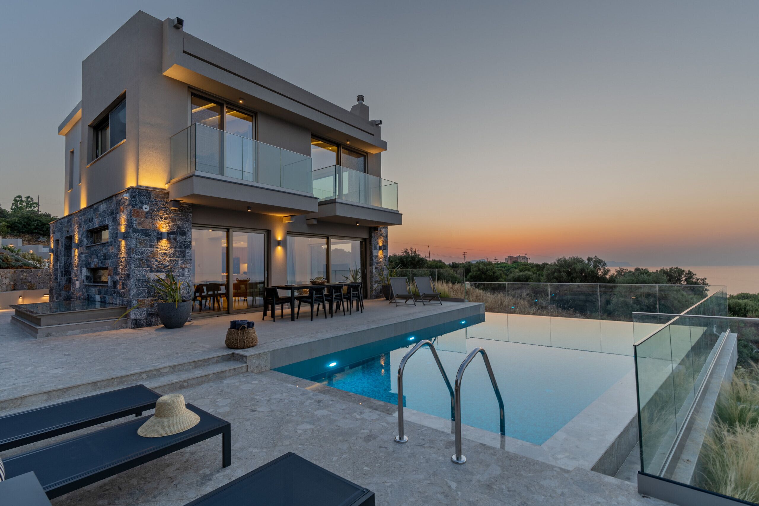 A modern two-story house with a swimming pool, outdoor furniture, and a view of the sunset.