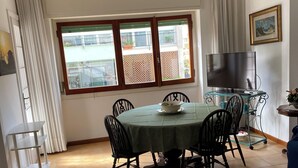 Un comedor con una mesa redonda, sillas, una araña, un televisor sobre un mueble y un ventanal con vistas a edificios.