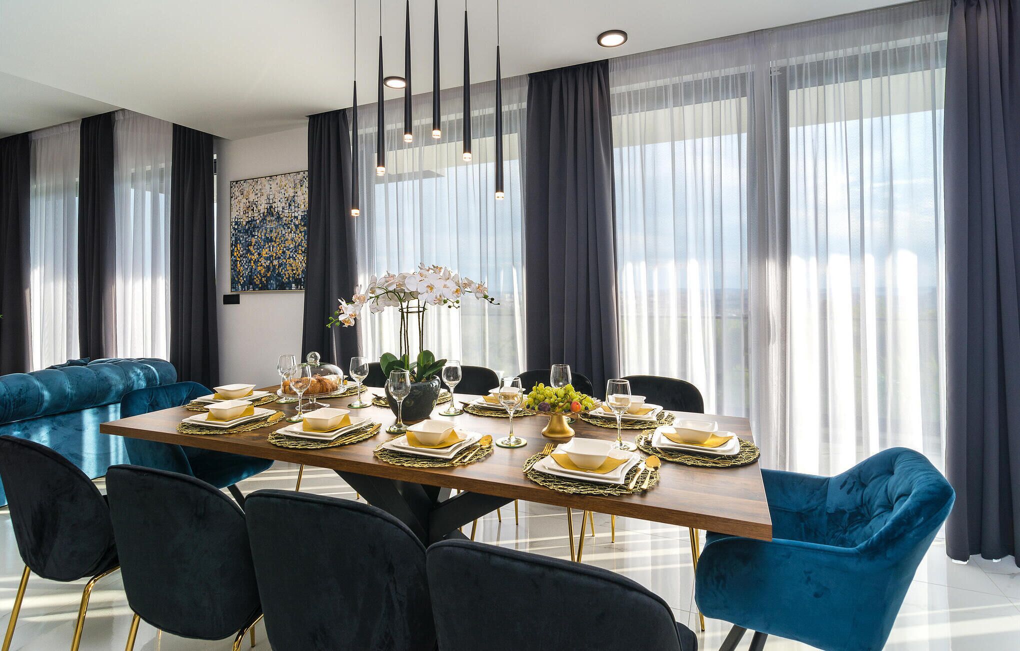 Un comedor con una larga mesa dispuesta para una comida, sillas de terciopelo azul, un jarrón de flores y una lámpara de araña moderna.