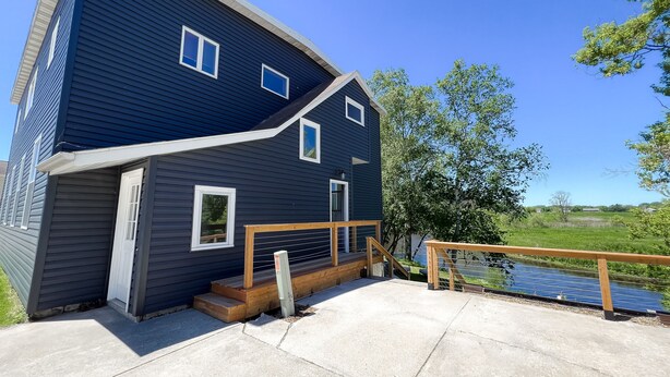 A two-story house with a dark blue exterior, a wooden deck, and a small wooden staircase leading to a porch. - Relax N Rentals Waterfront 3BR UP Algoma WI Kayaks Grill Fire Pit (Algoma)