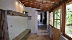 Un pasillo con techo de madera, un banco, un estante con plantas y una ventana que da a zonas verdes.