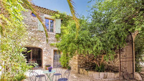 Edificio de piedra con un patio que incluye una mesa y sillas, rodeado de vegetación y una reja de metal.