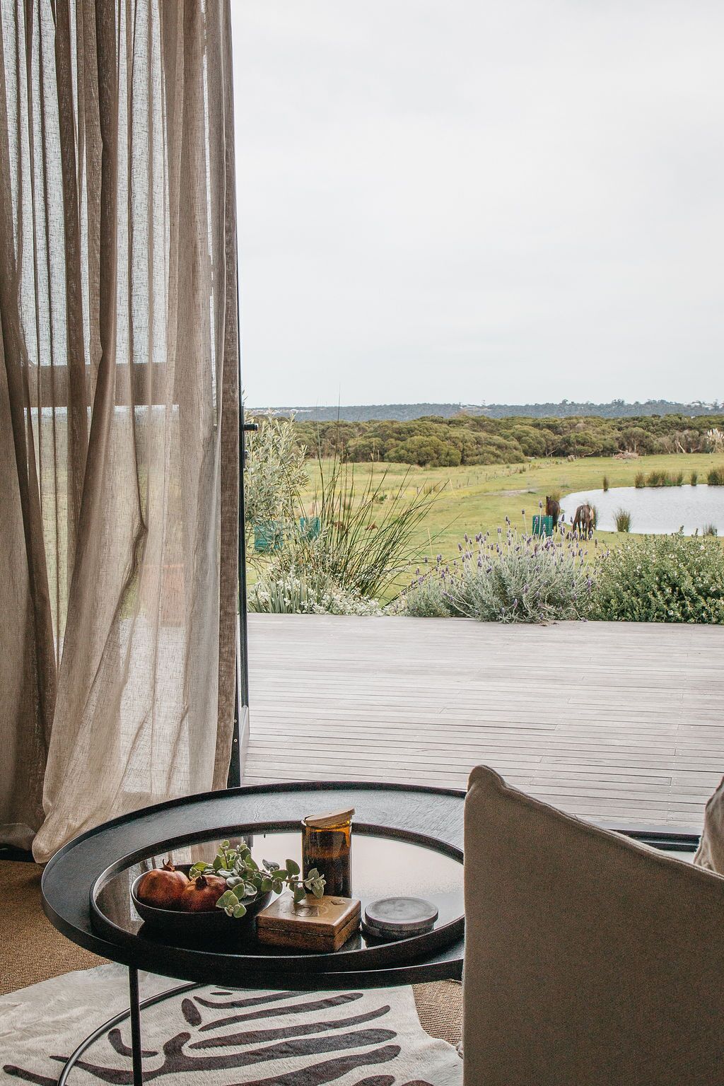 A room with a view of a lake and greenery, a round table with a bowl and candle, and a chair.