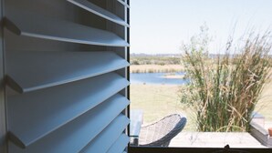 A view through a window with horizontal blinds, overlooking a grassy area with a body of water and some vegetation. - Majuba Country Lodge (Kalgan)