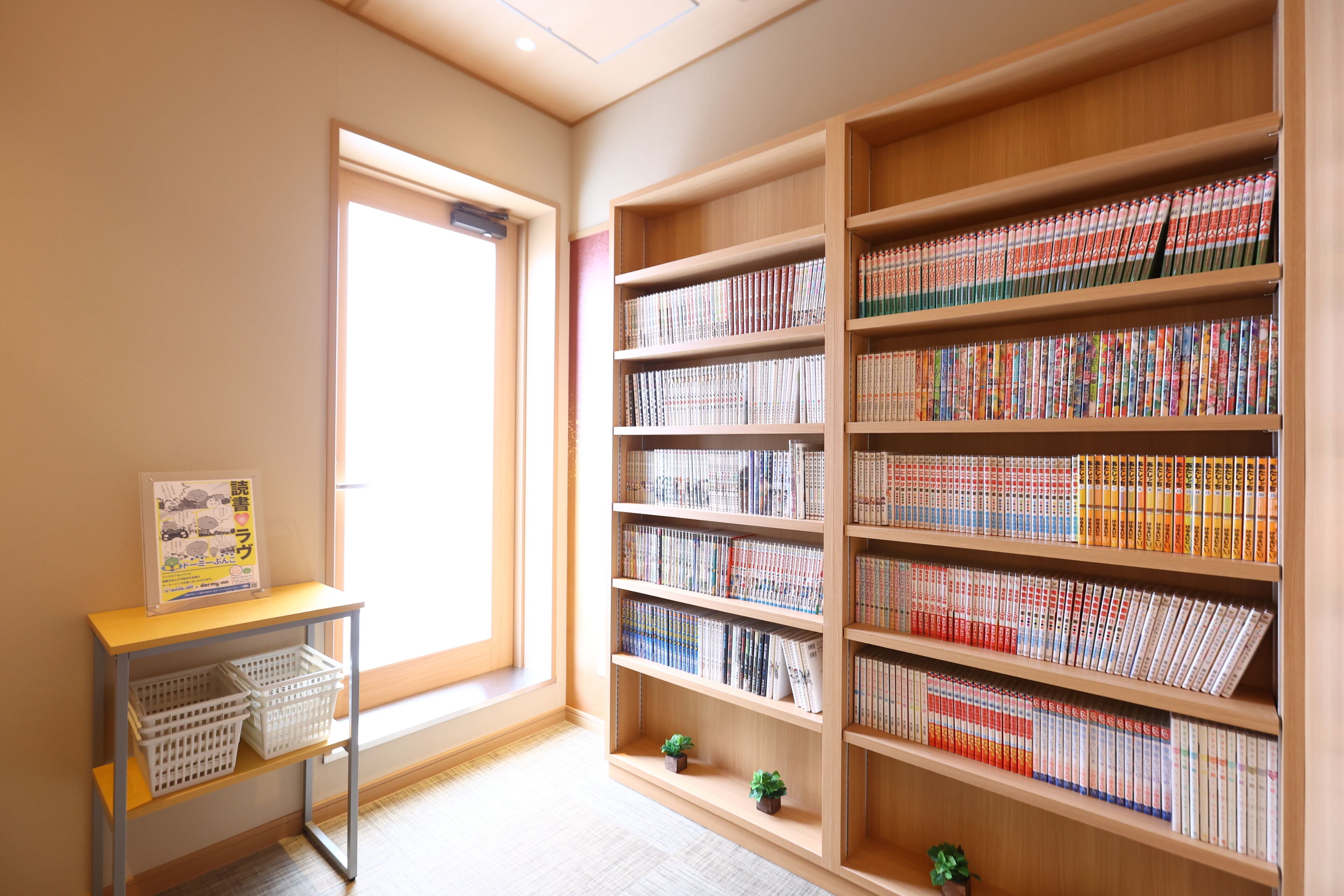 Una biblioteca con estanterías de madera llenas de libros, una pequeña mesa con cestas y un ventanal que proporciona luz natural.