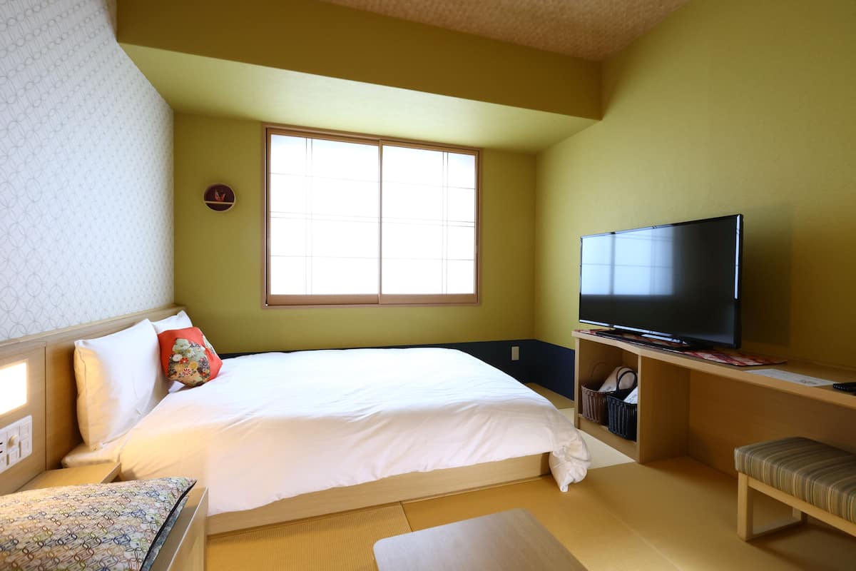 A hotel room with a bed, a television, a window, and a small seating area.