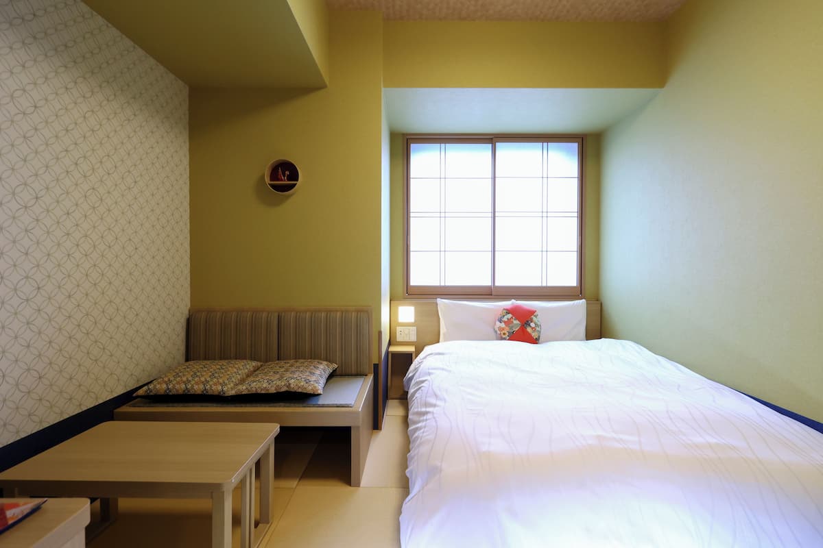 A hotel room with a bed, a bench, a bedside table, and a window.