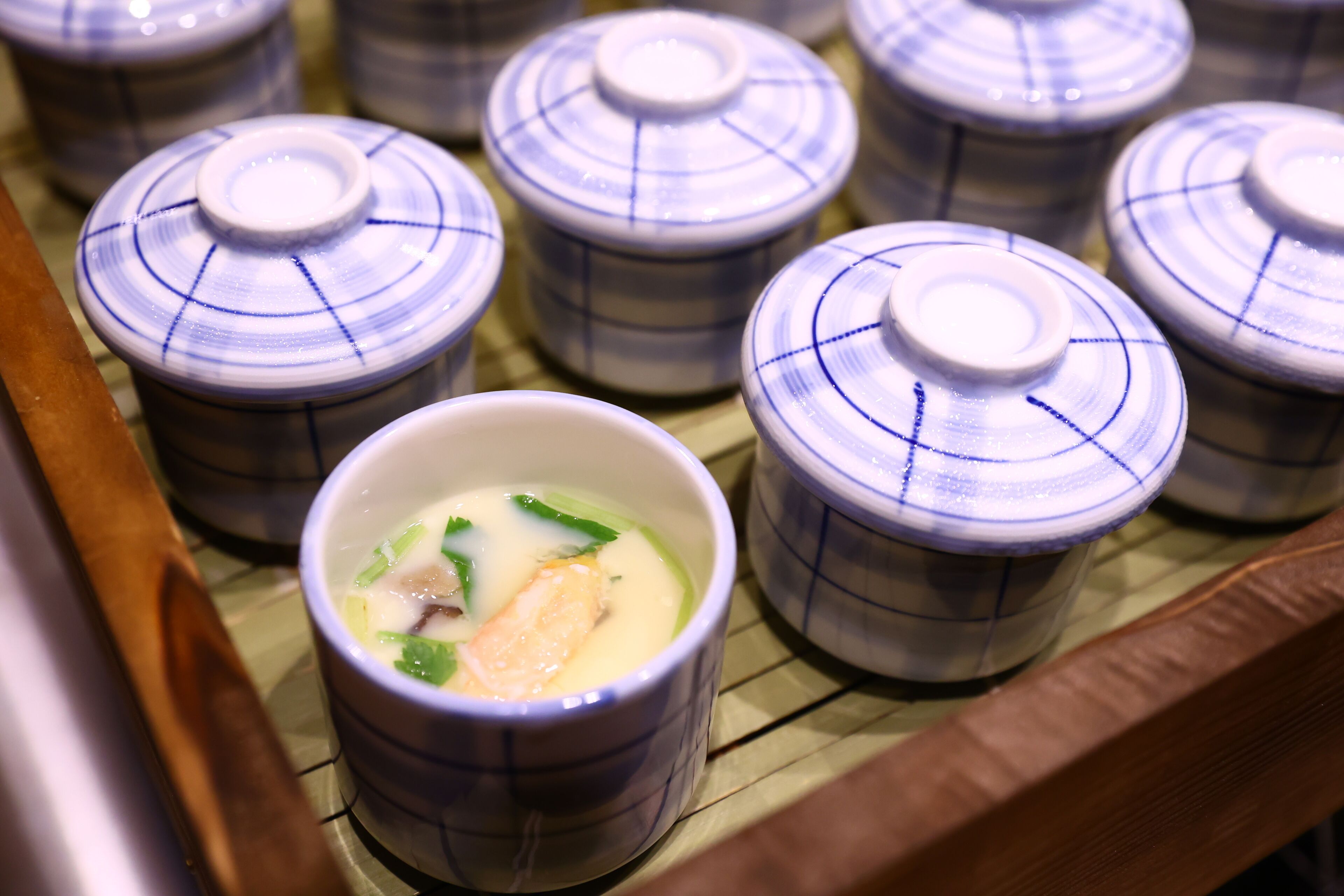 A row of ceramic bowls with lids, some containing food, placed on a bamboo mat.