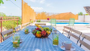 Una mesa con un frutero, vasos y una taza, ubicada al aire libre con una piscina y sillas al fondo.