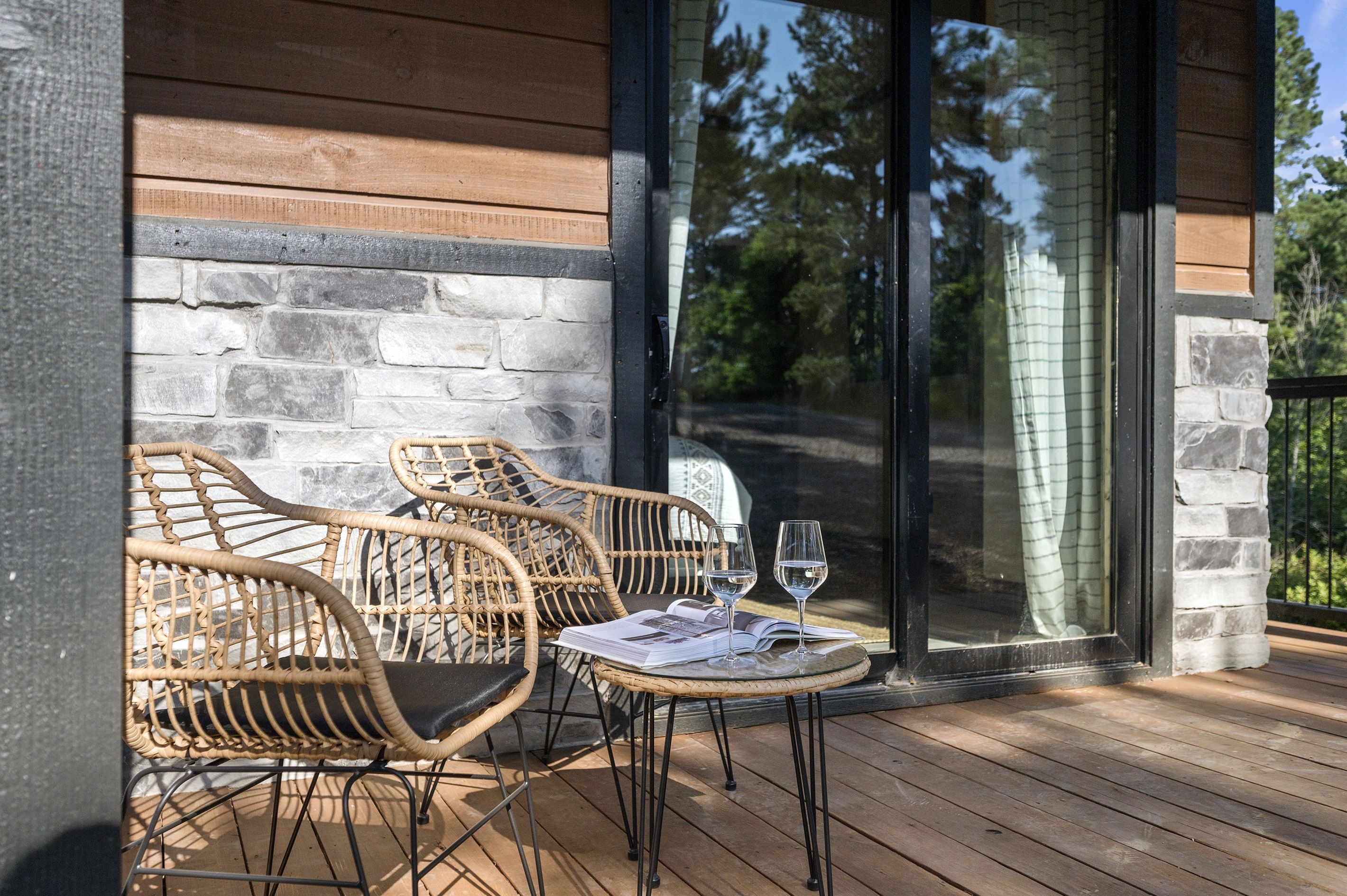 Une terrasse en bois avec des chaises et une table en osier, une porte vitrée et un mur en pierre.