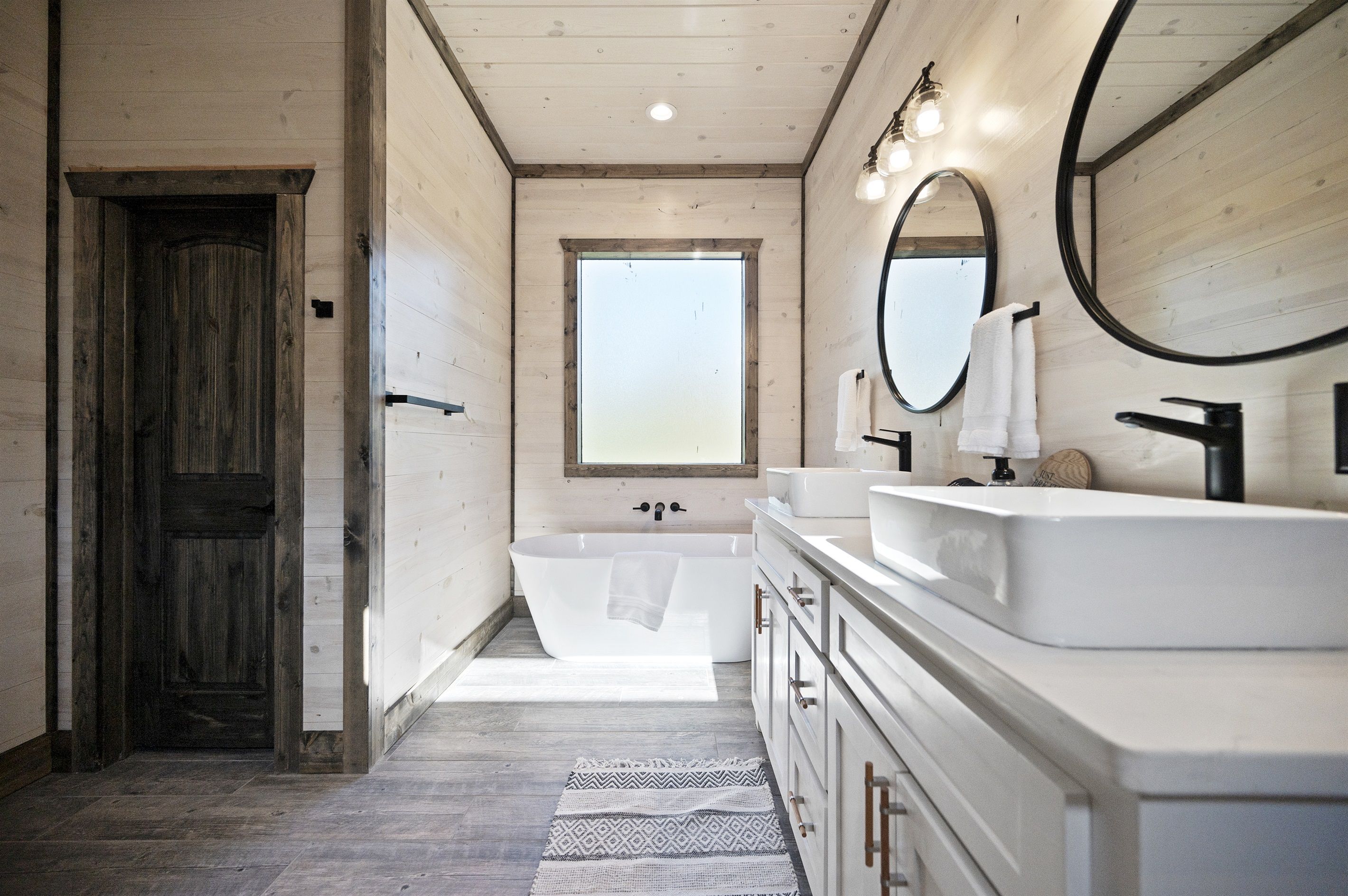 Une salle de bain moderne avec un intérieur en bois, une grande baignoire, deux lavabos et une fenêtre qui laisse entrer la lumière naturelle.