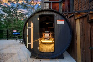A wooden barrel-shaped structure with a bench inside, a grill outside, and a warning sign.