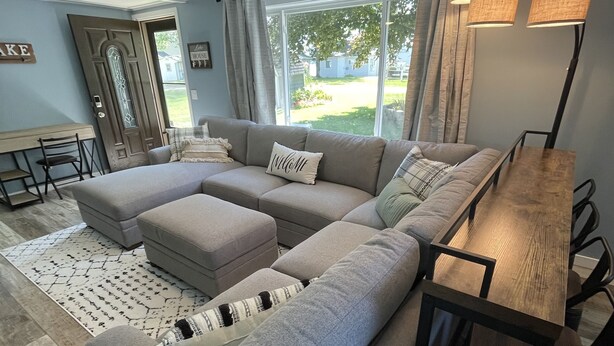 A modern living room with a sectional sofa, a wooden dining table, and a large window with curtains. - Peaceful lake retreat (Coldwater)