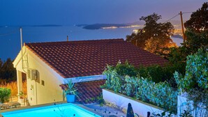 Et hus med swimmingpool, rødt tegltag og udsigt til vand i skumringen.
