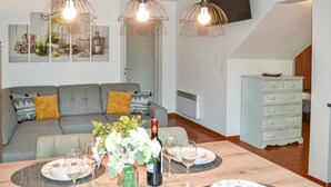 Ein modernes Wohnzimmer mit einem grauen Sofa, gemusterten Kissen, einem Esstisch, der mit Tellern, Gläsern und einer Weinflasche gedeckt ist. Über dem Tisch hängen drei Pendelleuchten und an der Wand ist ein Flachbildfernseher angebracht.