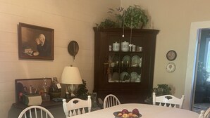 A dining room with a round table, white chairs, a fruit bowl, a lamp, a cabinet with dishes, and a painting on the wall.