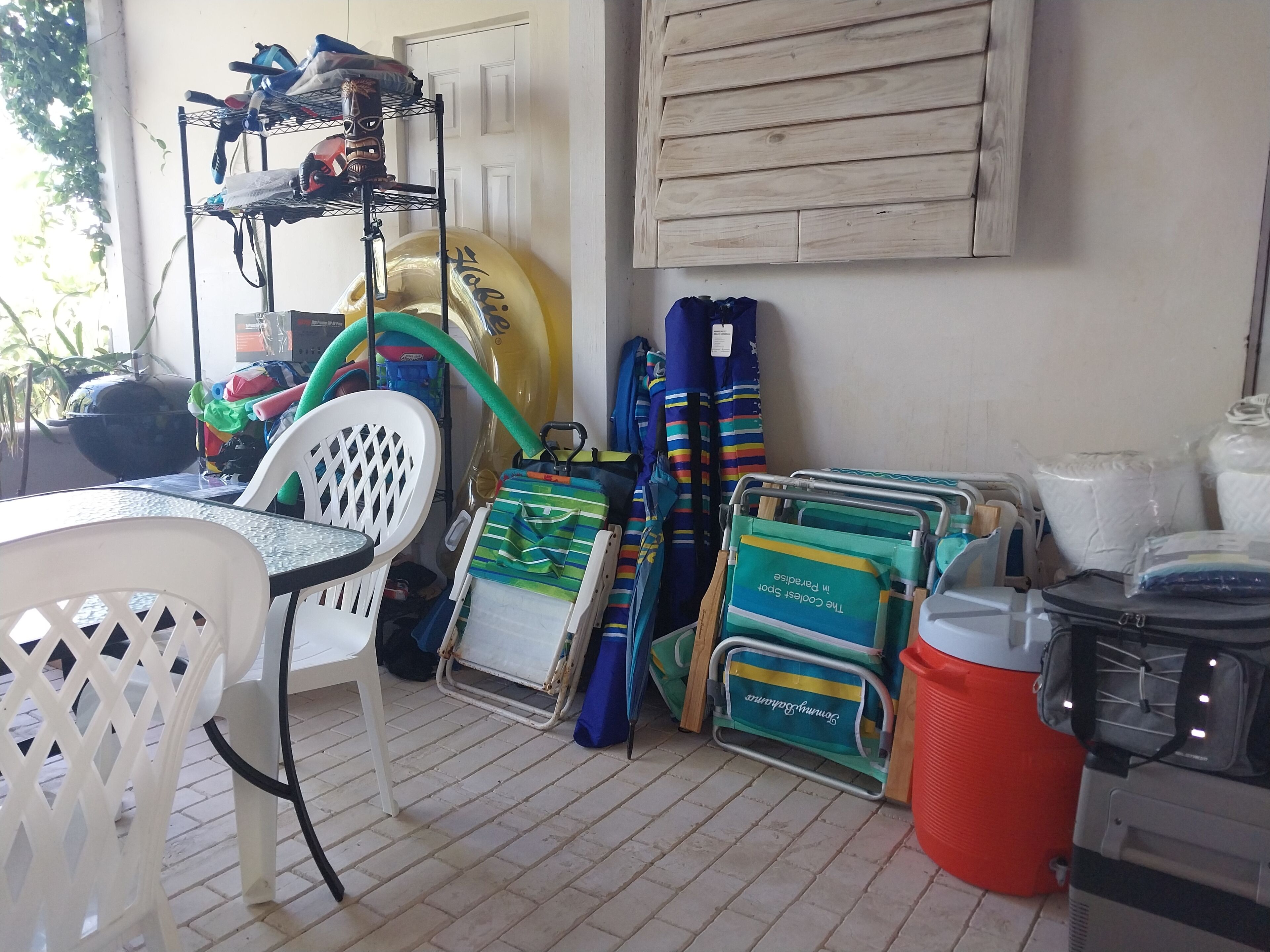 A room with a table, chairs, a chair with a mesh back, a shelf with items, a large yellow object, a blue and green bag, a cooler, and a wall with a wooden panel.
