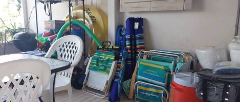 A room with a table, chairs, a chair with a mesh back, a shelf with items, a large yellow object, a blue and green bag, a cooler, and a wall with a wooden panel.