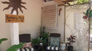 En uteplats med två fåtöljer i rotting, ett bord med en skål och ett ljus, en skylt med texten ”Sapodilla Villa” och en dekorativ solslinga.