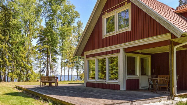 Une cabane en bois rouge, dotée de grandes fenêtres, d’une terrasse et d’un banc offrant une vue sur un lac.