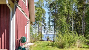 Un bâtiment rouge avec un porche en bois, une pompe à eau verte et une vue sur un lac entouré d’arbres.
