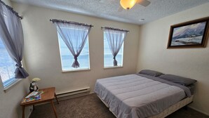 Une chambre à coucher avec un lit, deux fenêtres ornées de rideaux légers, une petite table avec une lampe et un tableau accroché au mur.