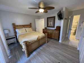 A bedroom with a wooden bed, a nightstand, a dresser, a ceiling fan, a television, and a door leading to a bathroom.