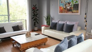 Una sala moderna con un sofá blanco, un sillón blanco y una mesa de centro de madera. Hay un cuadro en la pared y una planta en maceta.