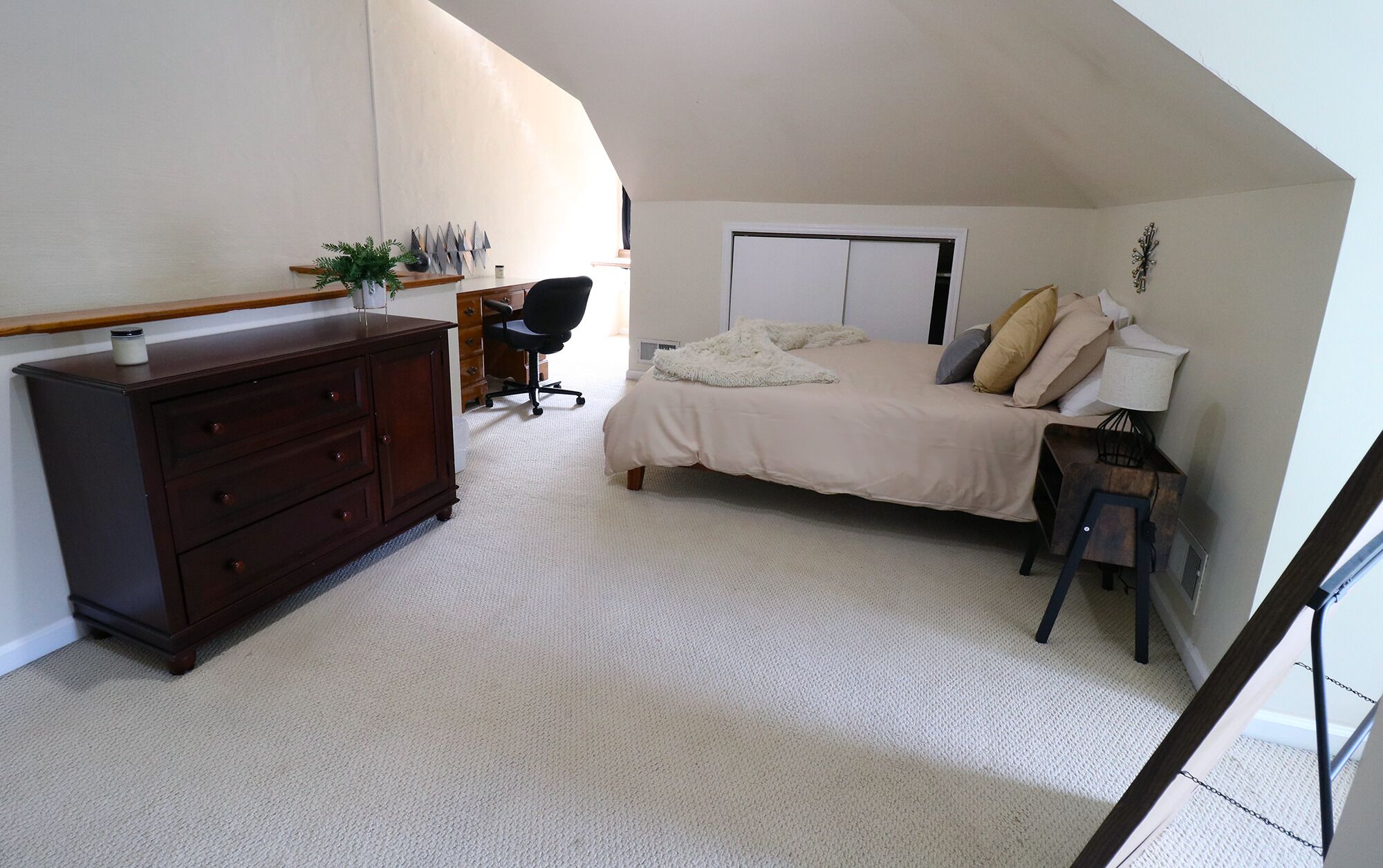 A bedroom with a bed, dresser, desk, chair, and a small table with a lamp.