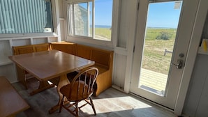 Une pièce avec une table et un banc en bois, de grandes fenêtres donnant sur la plage et une porte dotée d’une poignée en métal.