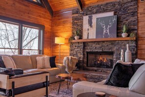 A cozy log cabin interior with a stone fireplace, leather sofas, and a large window.