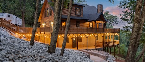 A log cabin with a stone pathway leading to a wooden deck, surrounded by trees.