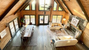 A spacious loft with a high ceiling, wooden beams, a dining area with a long table and chairs, a seating area with a sofa and armchairs, and large windows offering views of the outdoors. - Luxe Mountain getaway only 15 minutes from the city! (Greenville)