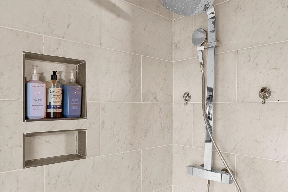 Et bruseområde med en håndholdt brusehoved og en vandhane fastgjort til væggen. Der er tre flasker shampoo og conditioner på en hylde.
