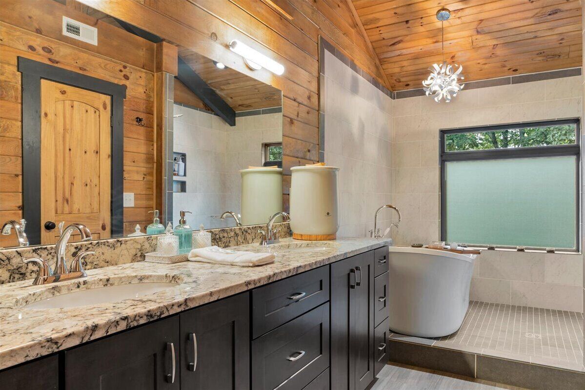 A modern bathroom with a wooden ceiling, a large mirror, a marble countertop, a bathtub, and a window with a view of trees.