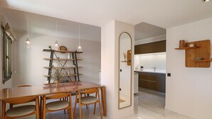 A modern dining area with a wooden table and chairs, a decorative shelf with plants, and a kitchen visible through an open doorway.