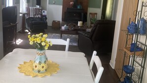 Ein Essbereich mit einem weißen Tisch, Stühlen und einem Blumenstrauß. Ein Kamin aus Backsteinen, ein Deckenventilator und ein Fernseher sind im Hintergrund zu sehen.